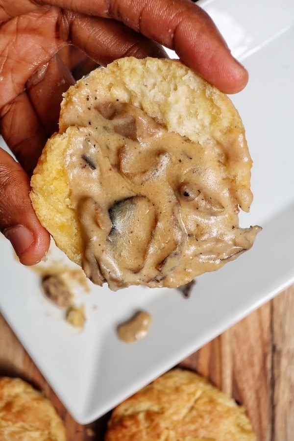 Hand holding half a vegan biscuit with mushroom gravy above a white plate